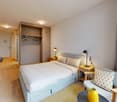 Spacious bedroom with a large bed, wooden wardrobe, bedside tables, and a comfortable chair. The room has large windows and a balcony or terrace area.