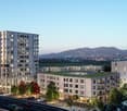 Multi-story apartment building with balconies, parking spaces, and green spaces. The building is surrounded by trees and has a view of the hills and mountains in the background.