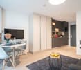 The image shows a modern and stylish studio apartment. The main living area features a round dining table with chairs, a TV mounted on the wall, and built-in kitchen cabinets and appliances, including a dishwasher. The apartment has a minimalist and conte