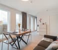 Spacious living room with large windows, wooden dining table, sofa, and storage cabinet. The room has a modern and minimalist design with neutral colors.