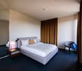 The image shows a modern and minimalist bedroom with a gray upholstered bed, white bedding, and brown curtains covering the window. There is a small side table with a lamp and a framed artwork on the wall. The room has a clean and simple design with a dar