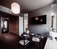 The image shows a modern and stylish hotel room. It has a large flat-screen TV mounted on the wall, a round dining table with two chairs, and a small side table with a lamp. The room has a dark color scheme with black and gray tones, creating a cozy and s