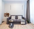Cozy living room with a gray sofa, decorative pillows, a wooden shelf, and a plant. The room has large windows and wooden flooring.