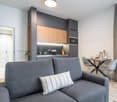 Modern studio apartment with open-concept layout. Gray sofa, dining table with chairs, and a kitchen with wood-tone cabinets and a built-in dishwasher. The space has a clean, minimalist design with white walls and hardwood flooring.