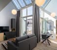 Spacious living room with high ceiling and large windows, providing ample natural light. The room features a gray sofa, a TV stand, and a dining table with chairs. The room also has a balcony or terrace accessible through the glass doors.