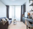 cozy grey sofa, large window with curtains, wooden floor, built-in entertainment unit, desk, and chair, floor lamp, open shelving with decor