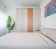 Bedroom with a bed, white walls, white curtains, a plant, a wardrobe, a picture frame, and a colorful painting on the wall.