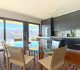 Modern kitchen with dark cabinets, glass dining table, and chairs. Large windows provide a scenic view of the city and mountains in the background.
