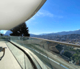 Spacious balcony with glass railing, offering a panoramic view of the city and surrounding mountains. The balcony appears to be part of a high-rise apartment or condominium unit.