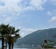 The image shows a scenic view of a lake surrounded by mountains and palm trees. The sky is partly cloudy, creating a picturesque atmosphere. There are some buildings and structures visible in the distance, suggesting a lakeside town or resort area.