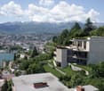 Modern multi-story building with a terrace overlooking a scenic mountain landscape and a lakeside town. The building is surrounded by lush greenery and has a panoramic view of the mountains and the lake below.