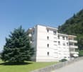 A multi-story apartment building with a white exterior, surrounded by trees and greenery. The building has several balconies and windows, and appears to be located in a mountainous, scenic area.