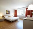 Spacious bedroom with hardwood flooring, large windows, white walls, a double bed, bedside tables, a dresser, and a floor lamp. The room also features red curtains and a framed artwork on the wall.