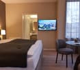 a bedroom with a bed, a remote control, a nightstand, a wall-mounted TV, a chair, a round table, a curtain, a pillow, a bathroom with a sink, and a lamp