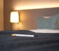 Bed with gray sheets, two pillows, lamp behind bed, chocolate wrapper on bed