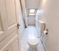 A small bathroom with white tiles, a toilet, a roll of toilet paper, a small window, and a light on the ceiling.