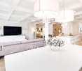 modern living room with white walls, wooden floor, large windows, white dining table, gray sofa, chandeliers, TV, kitchen with white cabinets, island with sink, flowers in vase