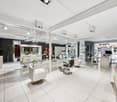 Modern salon with glass walls, white tiled floor, barber chair, shampoo station, makeup station, mirrors, lighting, and plants.