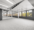 Large open office space with floor-to-ceiling windows, providing a view of the surrounding area. The space has a modern, minimalist design with gray carpeting, white walls, and recessed lighting. There are glass partitions dividing the space, creating a s