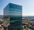Tall, modern glass office building with a sleek, angular design. The building has many floors and is surrounded by a cityscape with other buildings, streets, and green spaces visible in the background.