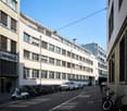 Modern office building with glass windows, multiple floors, and a sign reading 'Helvetic'. Street with parked cars and bicycles, and a pedestrian walking.