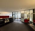 modern office space with glass walls, black and red furniture, carpeted floor, books on a shelf, a table with magazines, and a banner