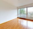 Empty room, hardwood floors, white walls, sliding doors leading to balcony, windows with view of trees and house, wall mounted light, power outlets on the wall.