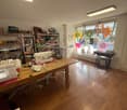 A room with a wooden table, sewing machines, shelves with clothes, a mirror, and a window with decorations.