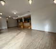 Large open space with hardwood-style tile flooring, white walls, and recessed lighting fixtures. The room appears to be a living or dining area with a kitchen visible in the background.