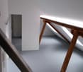 The image shows a room with white walls and a wooden table or desk with a triangular support structure. There is a white cabinet or shelving unit visible on the left side of the room. The room appears to be in a minimalist or modern style.