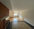 The image shows an empty room with a brick wall on one side and a tiled floor. The room has a hardwood floor and white walls, and there is a window visible. The room appears to be a living or dining area.