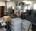 Storage area with several plastic bins, a cart, shelves, boxes, and printing equipment.