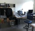 An office with a desk, chair, computer, printer, shelves with storage boxes, papers and bags.