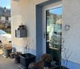 Concrete facade, gray mailbox mounted on the wall, entrance door with glass panel, multiple potted plants on the pathway