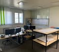 The image shows an office space with multiple desks, computers, and office equipment. The room has large windows, a whiteboard, and shelves with books and documents. The overall layout and furnishings suggest this is a professional work environment.