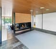 wooden ceiling, fireplace, black fireplace hearth, white rug, white walls, large windows, glass wall, chairs, brown tile floor