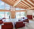 Spacious living room with high wooden ceiling, large windows offering scenic mountain views, leather sofas and armchairs, glass coffee tables, and a fireplace