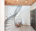 The image shows the interior of a building with a spiral staircase made of concrete and metal railings. The ceiling has a wooden design with recessed lighting. There is a fireplace visible in the corner of the room. The walls are white and the floor is ma