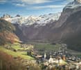 Mountain village with snow-covered mountains, a large church, and a clear blue sky with clouds.