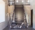 Metal staircase with open treads, leading to an upper level. The stairs have a modern, industrial design with a metal frame and glass railings. The space appears to be a loft or converted industrial space, with exposed concrete walls and artwork on the wa