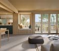 Open-concept living space with kitchen, dining area, and living room. Large windows and sliding glass doors leading to a balcony or terrace with views of the surrounding greenery. Wooden ceiling, neutral color palette, and modern furnishings.