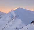 Snow-covered mountain peaks illuminated by a warm sunset glow, with rocky outcrops.