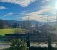View from a balcony or window, showing a large green field, trees, a house, mountains, and a clear blue sky with some clouds.