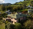Large two-story house with a red tile roof, surrounded by lush green hills and trees. The house has a garage and parking space visible. The landscape features rolling hills, trees, and other residential buildings in the background.