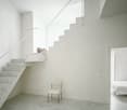 White marble staircase, white walls, open doorway leading to bedroom with a bed, small white chair, window with pillows on window sill.