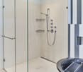 Large glass shower enclosure, multiple shower heads, built-in shelves, modern bathroom fixtures, and a dishwasher visible in the background.