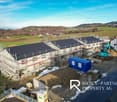Aerial view of a construction site with several houses under construction. Some houses have metal scaffolding, others have roofing materials installed. In the background are a tractor and various construction vehicles. The site is surrounded by rural gree