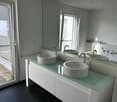 Modern bathroom with white vanity, glass countertop, vessel sinks, and faucets. Large windows provide natural light and access to a balcony or terrace.