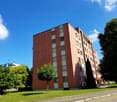 Red building, many windows, some with balconies, located on the grassy area, trees and street in front