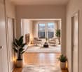 bright corridor with wooden flooring, living room with fireplace and balcony, white walls and ceiling, white furniture, large window with curtains, two armchairs, coffee table, potted plants